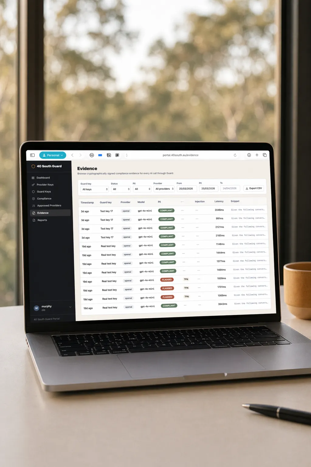 Close-up of a laptop on a wooden table showing the 40 South Guard evidence dashboard with compliance status badges; Australian bush visible through the window behind.